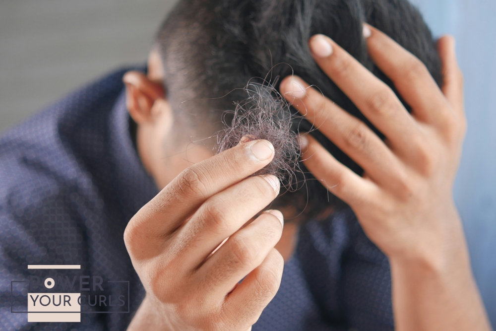 A man is out of focus in the background, holding his head with one hand. On the foreground is his other hand holding up a clump of what looks to be his own hair.