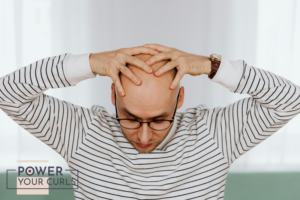 A man holding the top of his bald head with both hands, he is looking down with a sullen expression on his face.