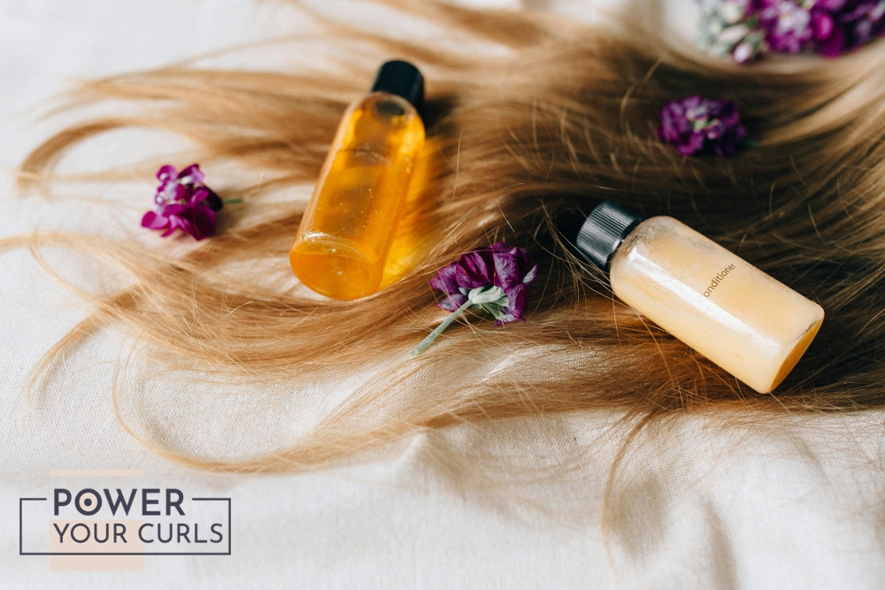 Two small bottles of shampoo and conditioner laid out onto soft, silky hair on a white sheet.