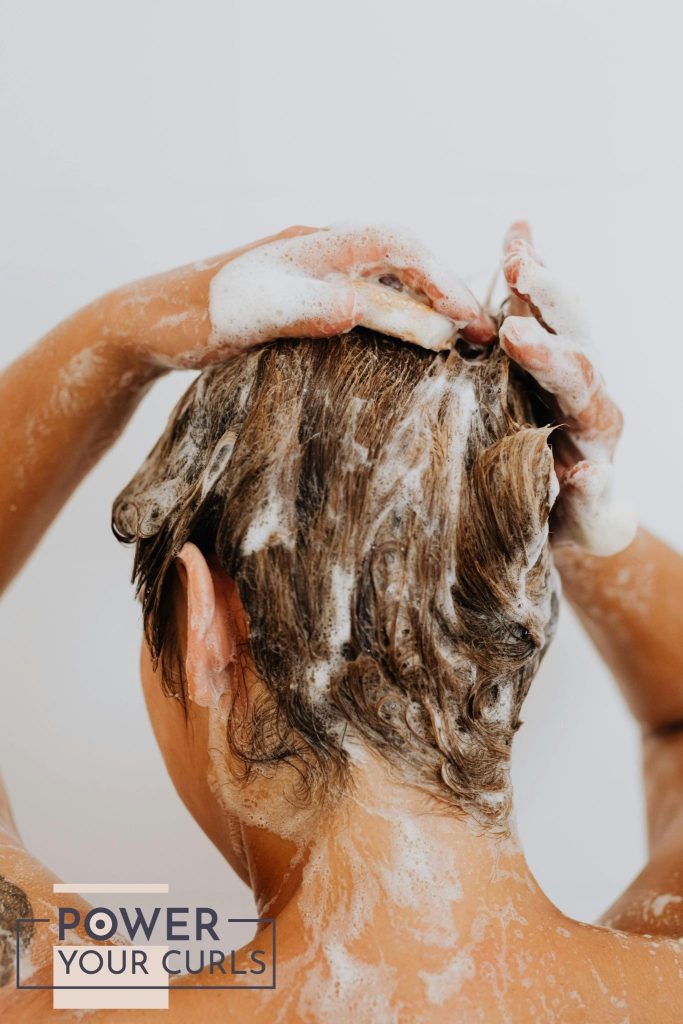 A photo of a woman, taken from behind, washing her hair with shampoo.