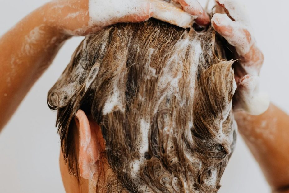 A photo of a woman, taken from behind, washing her hair with shampoo.