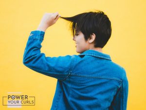 A woman with short hair playfully tugging at her bangs. She is wearing a jean jacket and is standing in front of a yellow background.