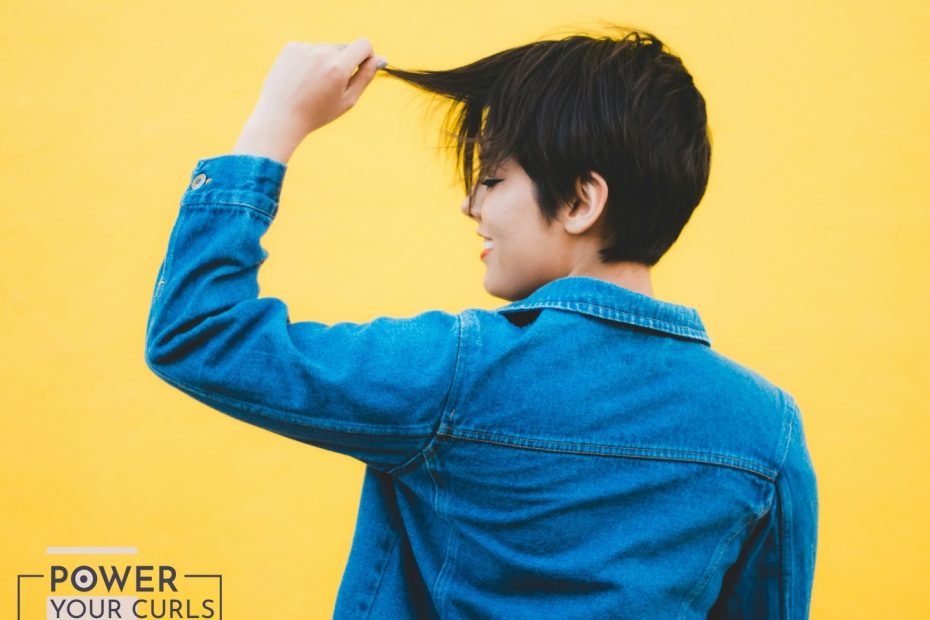 A woman with short hair playfully tugging at her bangs. She is wearing a jean jacket and is standing in front of a yellow background.