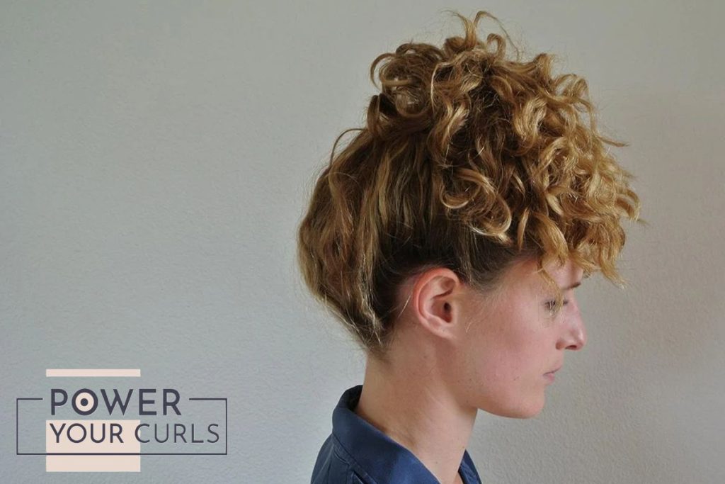 Image of a woman with blonde hair and darker roots, sporting a high pineapple hairdo for both every day and sleeping purposes while wearing a blue collared shirt