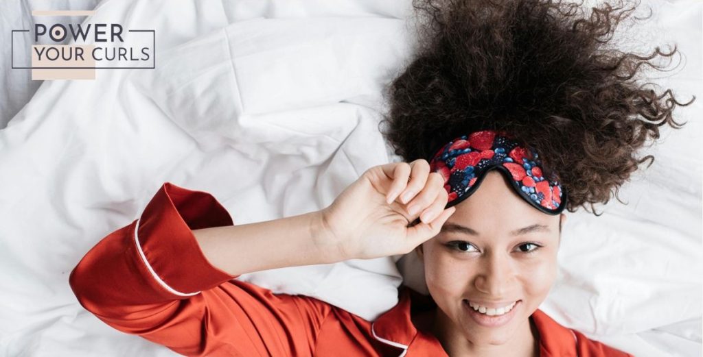 Image of a woman with dark curly hair laying down on the white seehst of a bed while wearing a red sleep shirt and a red-and-blue eyemask 
