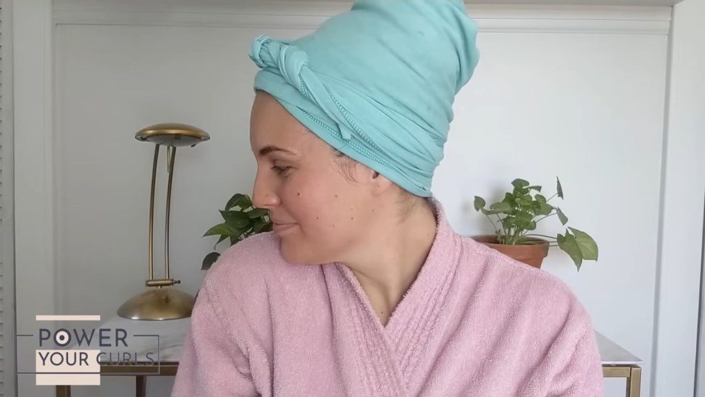 Image of a woman wearing a pink bathrobe and a blue t-shirt wrapped around her hair while standing in front of a while table with two potted plants and a gold lamp