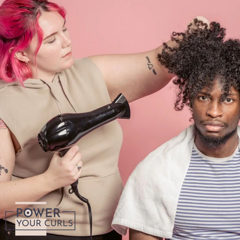 Why Is My Curly Hair Dry? Causes, Signs, and How to Fix It 2 Image of a woman with pink hair while wearing a beige sleeveless hoodie is blowing a man's curly hair dry with a blowdryer while wearing a blue-and-white stripped shirt and a white towel on his shoulder in front of a pink background